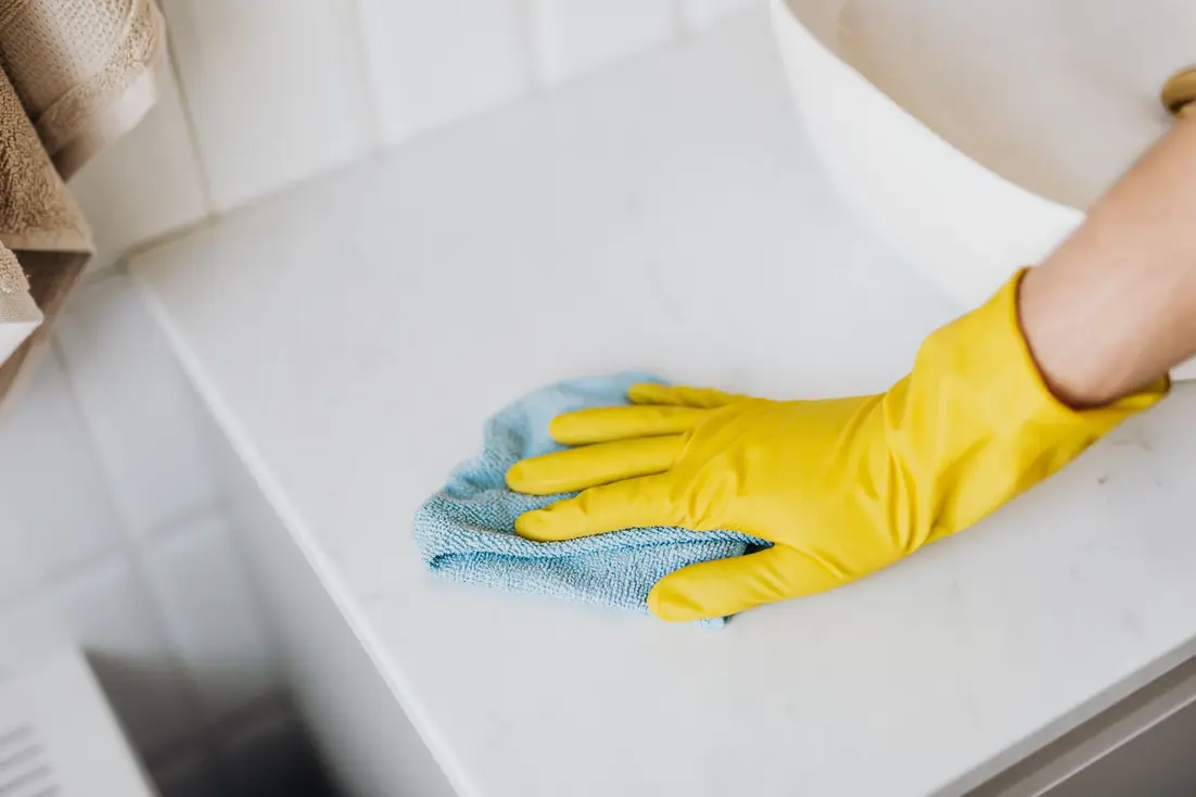 A hand in a yellow glove wipes a bathroom vanity with a blue cloth.